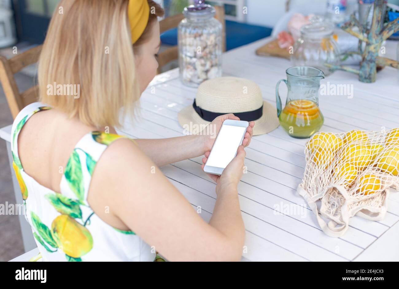 Ein blondes Mädchen sitzt an einem weißen Tisch, hält ein weißes Smartphone in den Händen, daneben ist ein Krug mit Limonade und Zitronen Stockfoto