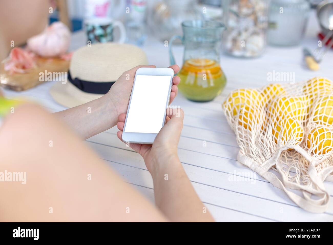 Hände eines Mädchens, das ein weißes Smartphone auf einem weißen Holztisch hält, neben gelben Zitronen in einer Tasche. Blick von oben. Stockfoto