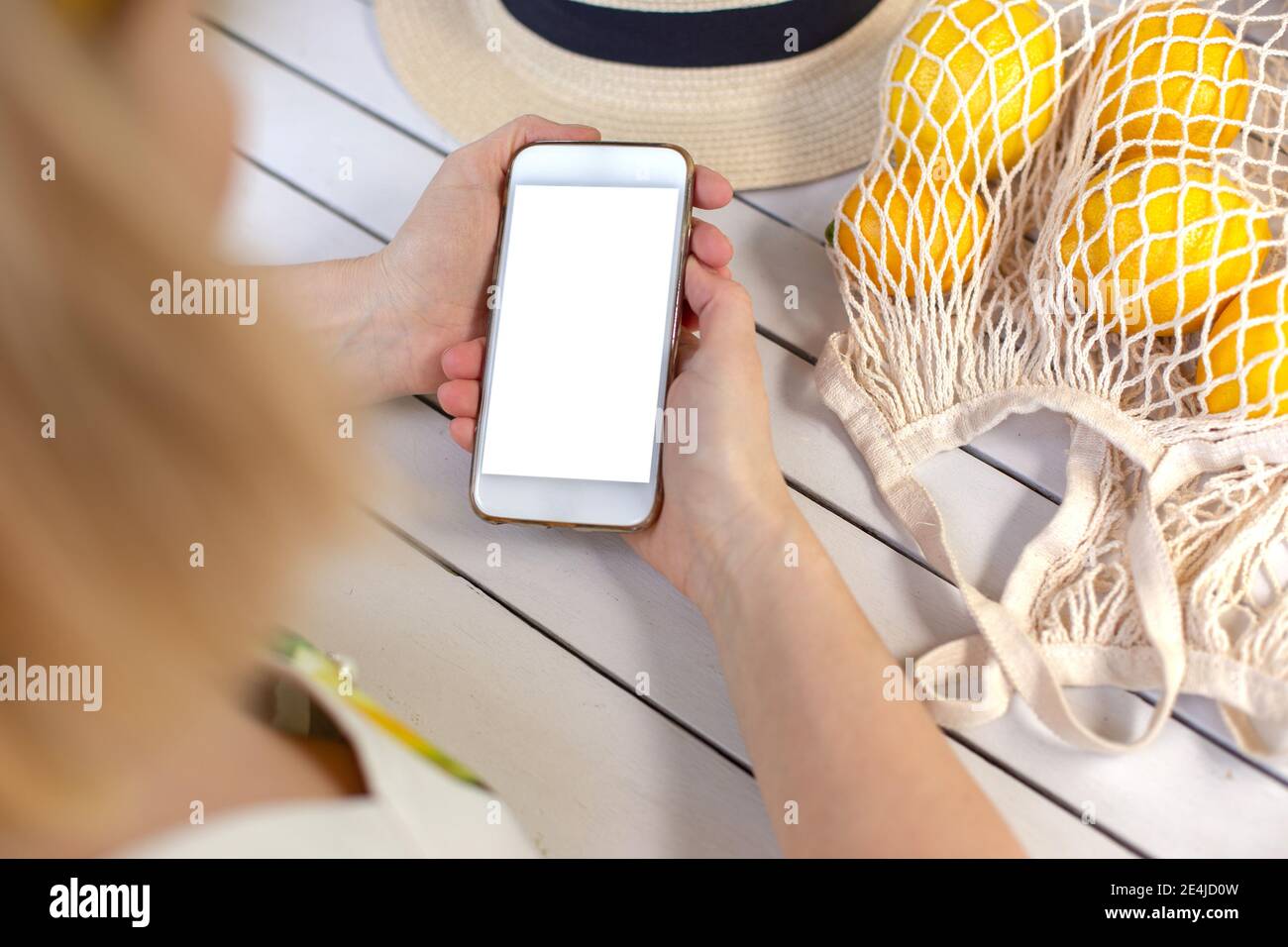 Hände eines Mädchens, das ein weißes Smartphone auf einem weißen Holztisch hält, neben gelben Zitronen in einer Tasche. Blick von oben. Stockfoto