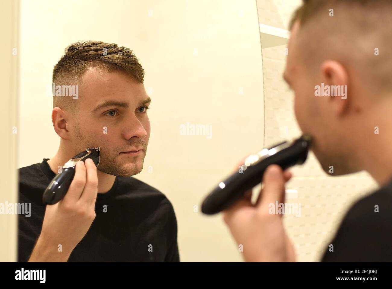 Junger Mann rasieren mit Trimmer und Blick in den Spiegel Im Badezimmer Stockfoto