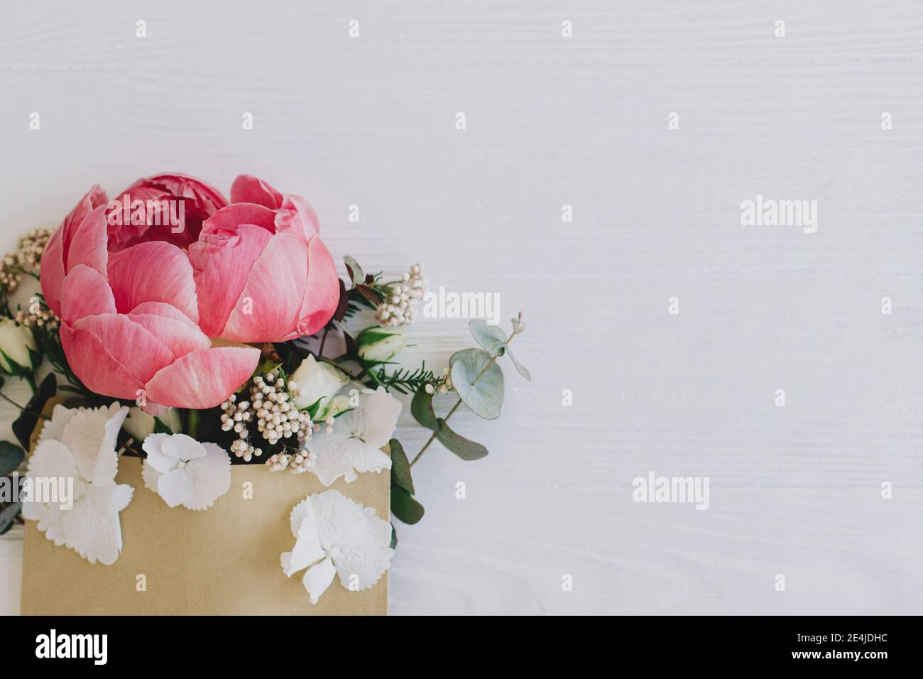 Pfingstrose, weiße Blumen und Eukalyptus wachsen aus Bastelhülle auf weißem Holz, flache Laie mit Platz für Text. Kreative Blumengrußkarte oder senden Stockfoto