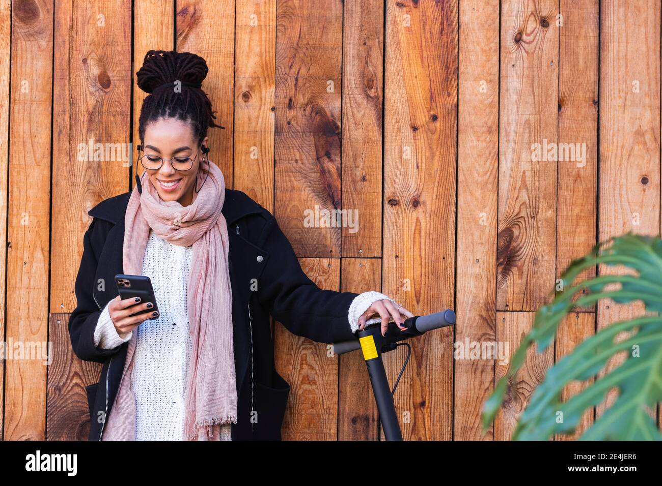 Schöne weibliche Millennial mit Smartphone von Elektro-Push-Roller Gegen Holzwand Stockfoto