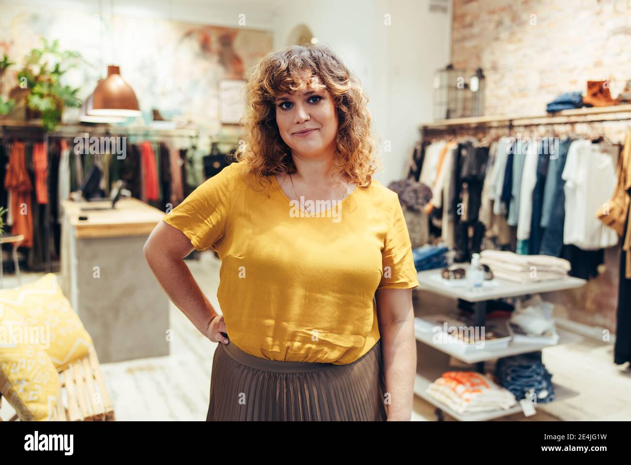 Porträt einer schönen Frau, die in einem Modehaus steht und mit der Hand auf der Hüfte auf die Kamera schaut. Selbstbewusster Boutique-Besitzer. Stockfoto