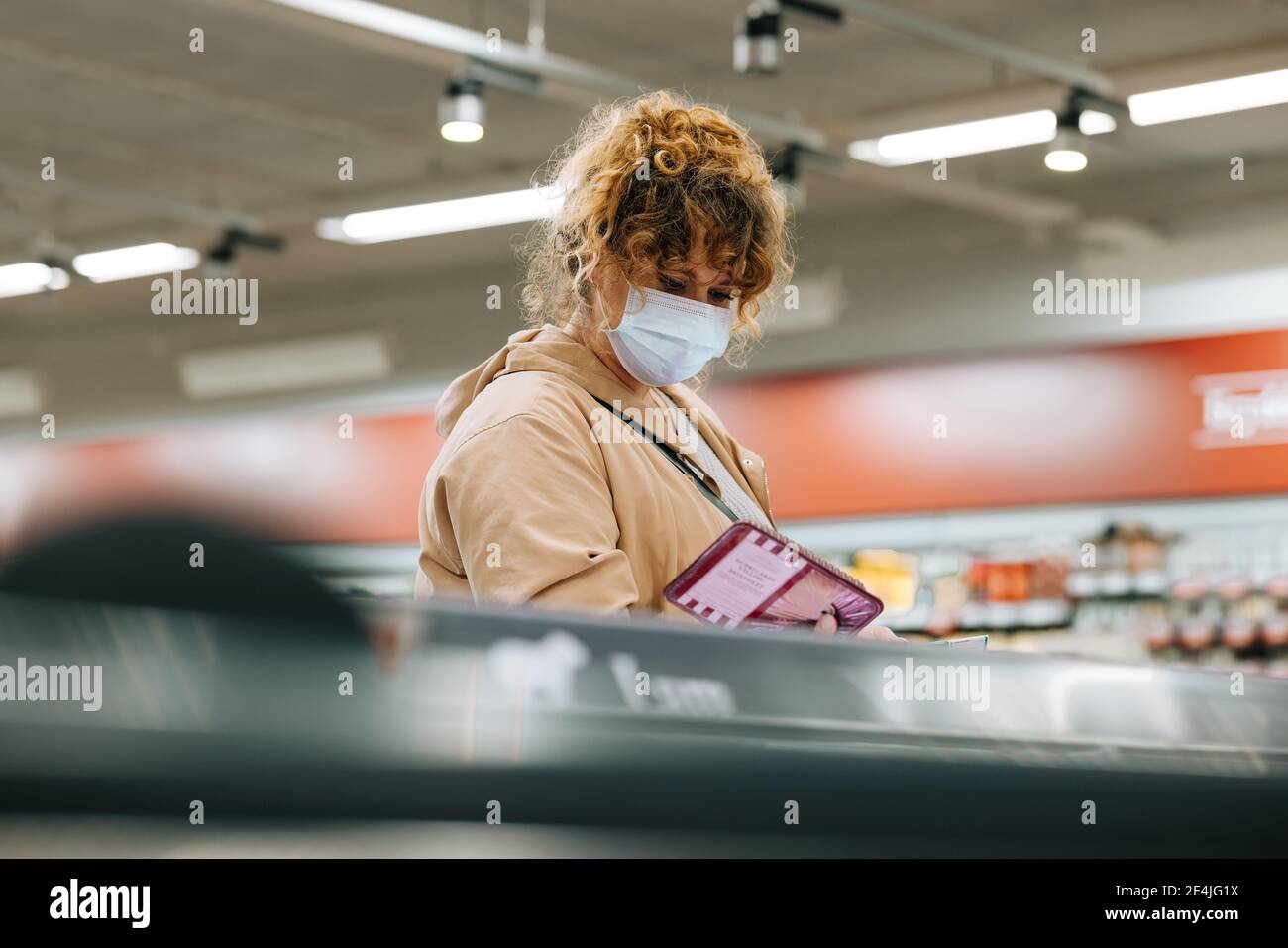 Frau mit Gesichtsmaske Einkaufen im Supermarkt. Weibliche Kundin trägt Schutzgesichtsbemask Blick auf Produkte im Supermarkt zu kaufen. Stockfoto