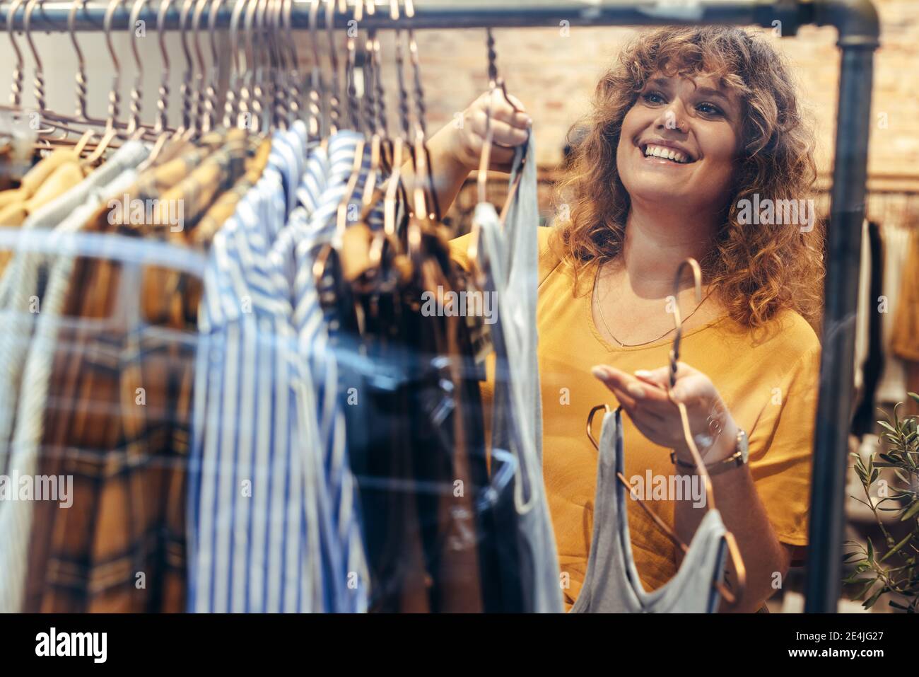 Glückliche weibliche Kundin, die sich für ein Kleid im Geschäft entscheidet. Frau mit mittlerem Erwachsenen, die in der Boutique steht und Kleider ansieht, die auf dem Regal hängen. Stockfoto