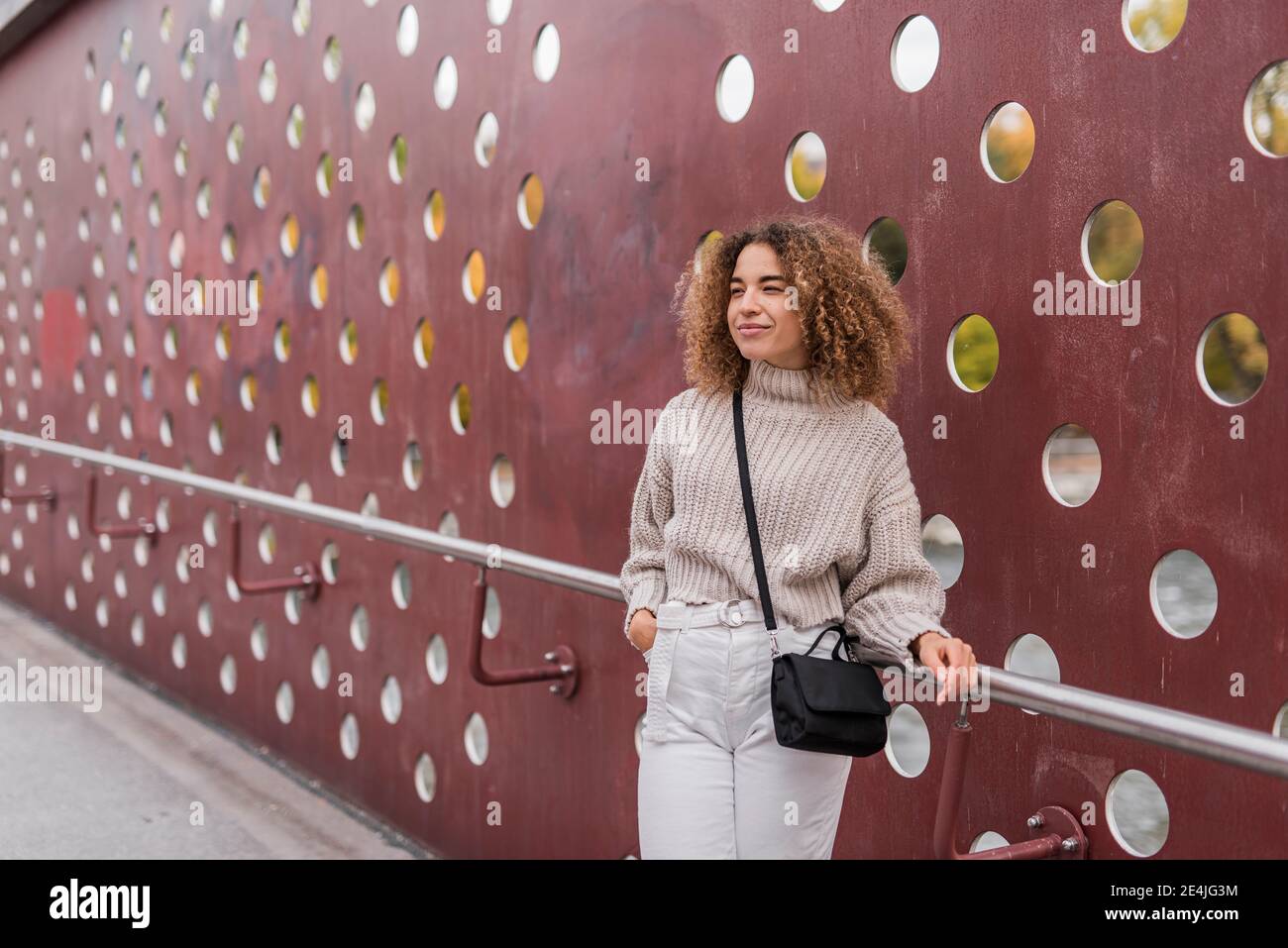 Schöne junge blonde Frau, die mit der Hand in der Tasche steht, während sie sich auf einem Geländer an einer metallischen Wand lehnt Stockfoto