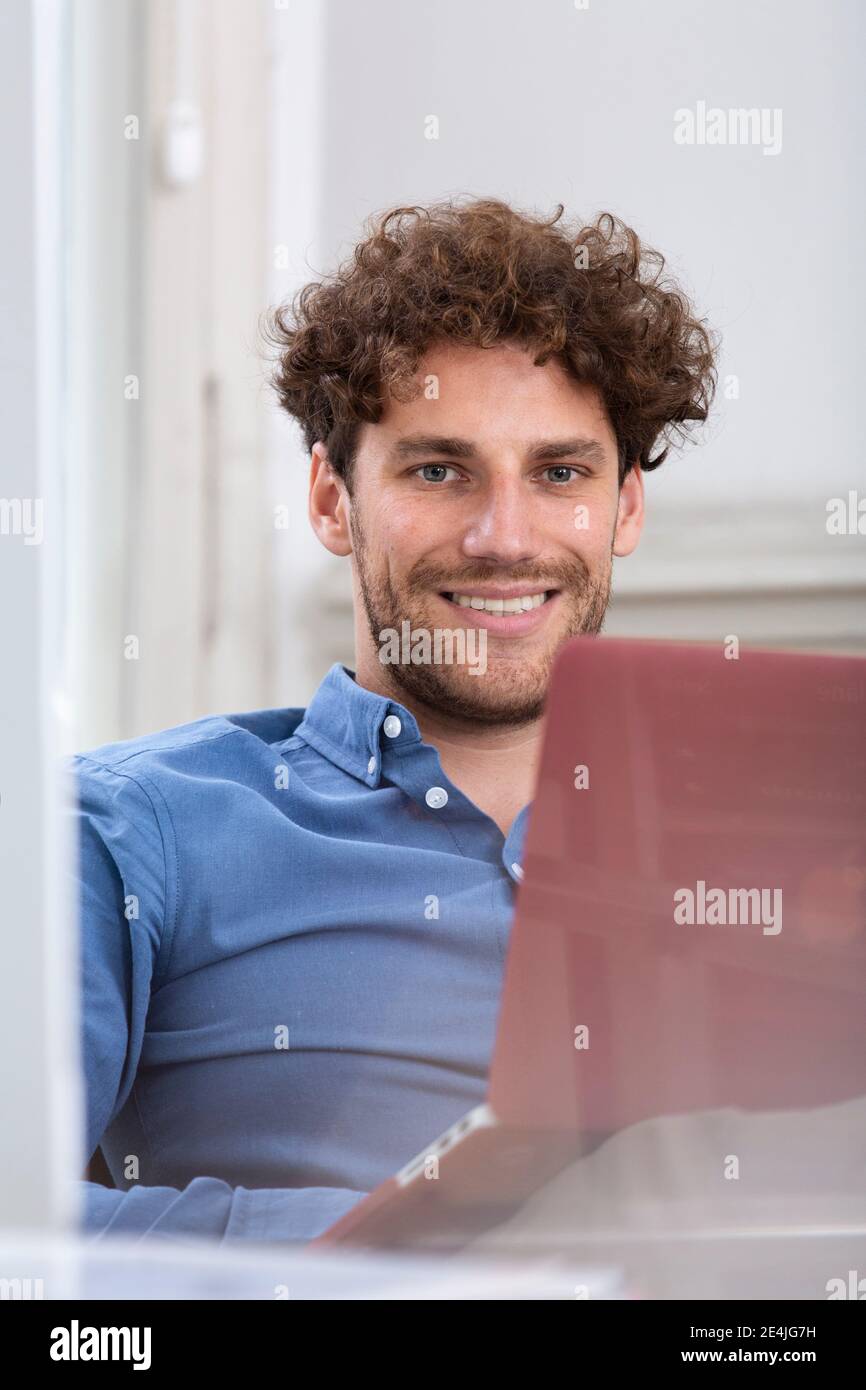 Männlicher Profi lächelt, während er mit Laptop in der Bürokabine sitzt Stockfoto Männlicher Profi lächelt, während er mit Laptop in der Bürokabine sitzt Stockfoto