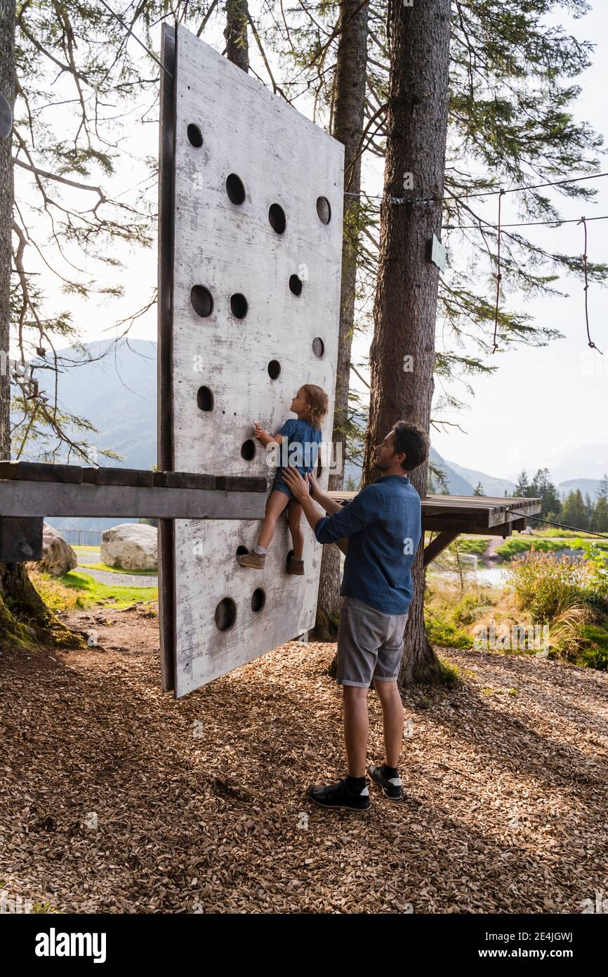 Vater unterstützt kleine Tochter versuchen, auf der Spitze zu klettern Kleine Waldkletterwand Stockfoto