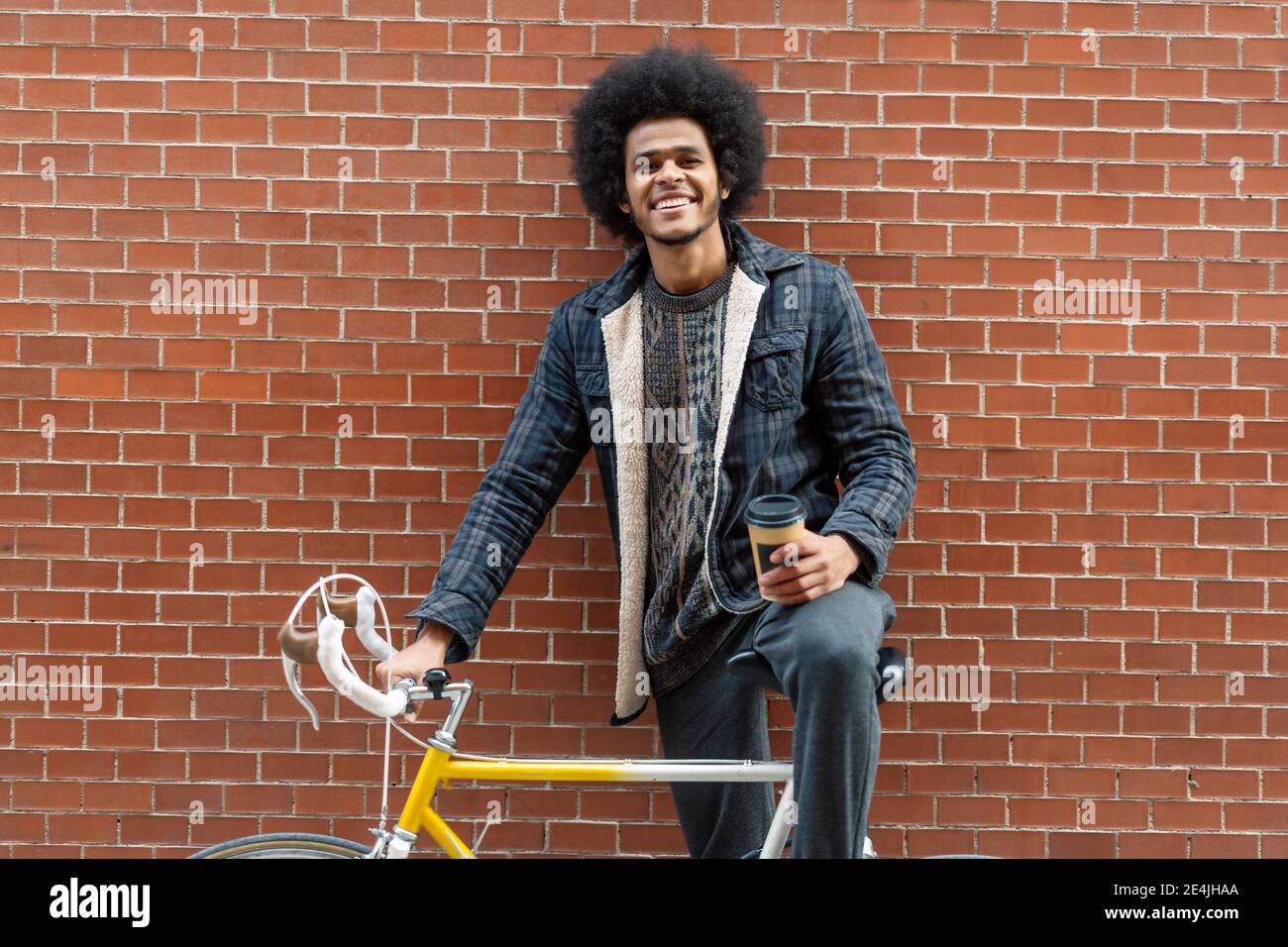 Junger Mann hält Kaffee, während er mit dem Fahrrad gegen Backstein steht Wand Stockfoto