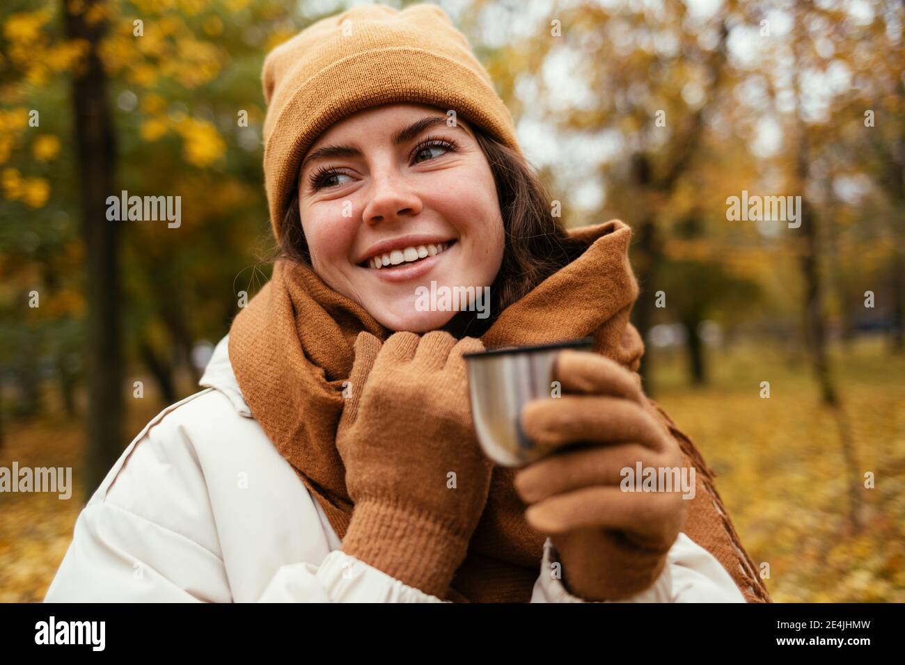 Glückliche junge Frau, die wegschaut, während sie isolierte Tasse in hält Herbstpark Stockfoto