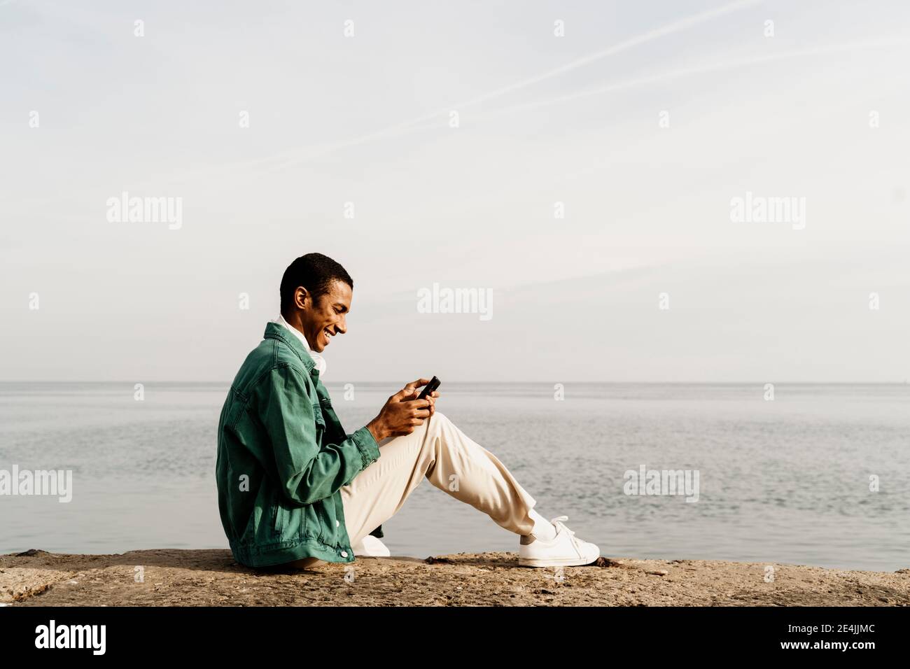 Glücklicher Mann SMS-Nachrichten über Smartphone, während sitzen auf pier am Himmel und am Meer Stockfoto