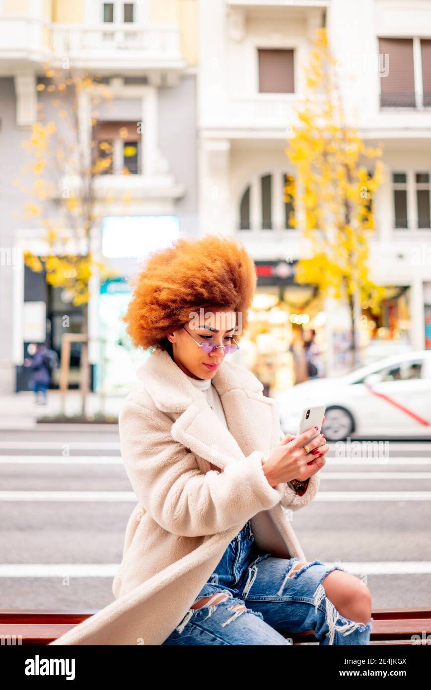 Frau mit afro blonden Haaren mit Smartphone während des Sitzens Auf der Bank in der Stadt Stockfoto