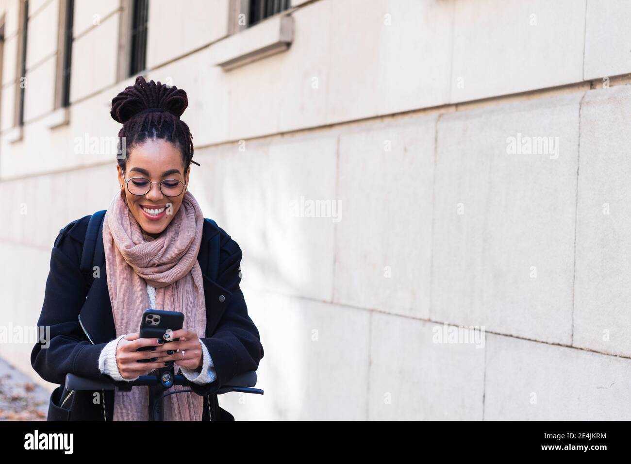 Lächelnd junge Frau Millennial mit Smartphone, während sich auf Elektroroller durch Gebäude schieben Stockfoto