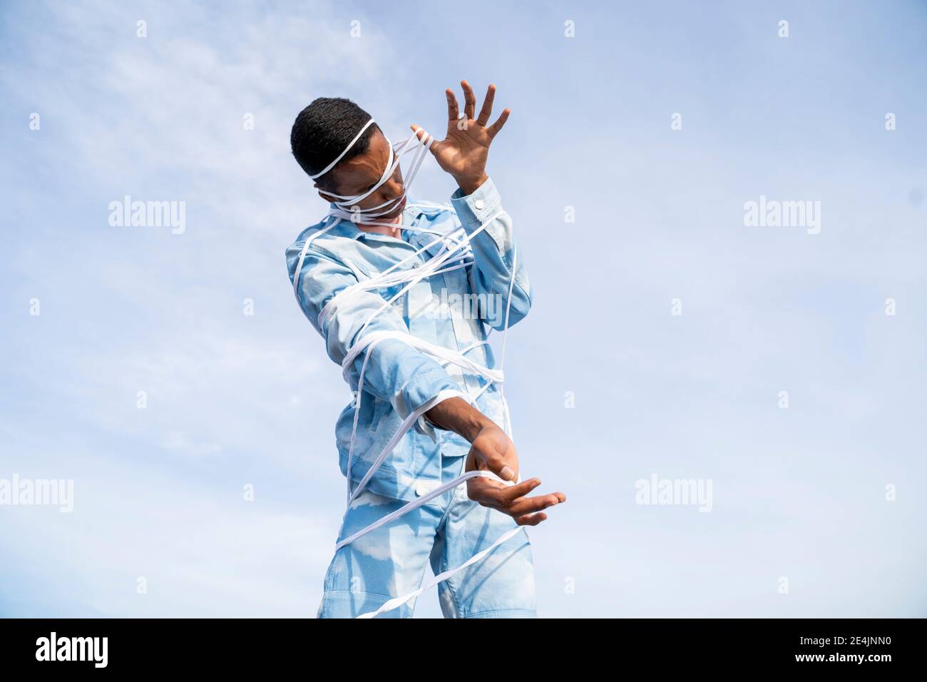 Junger Mann trägt Wolke gemustert Denim Outfit kämpfen, um zu bekommen Frei vom Seil, während man gegen den Himmel steht Stockfoto