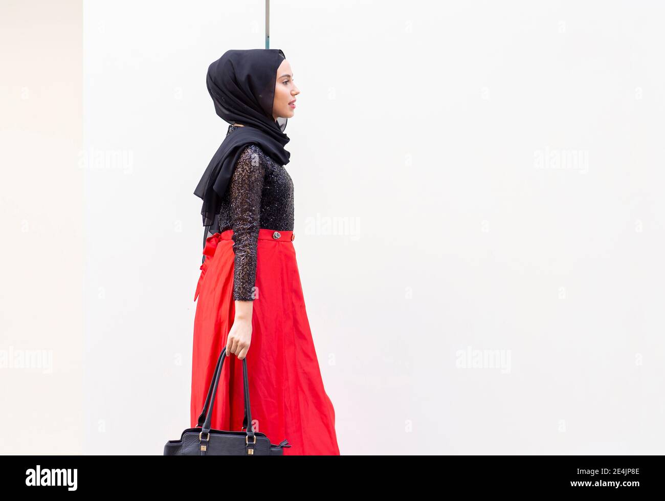 Junge schöne Frau trägt Hijab und langen Rock zu Fuß vor weiß Wand mit Handtasche in der Hand Stockfoto