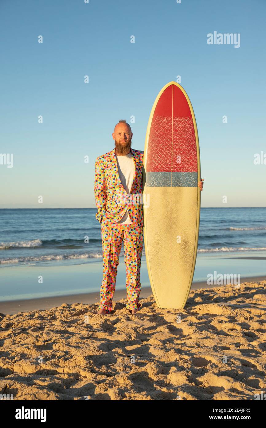 Modischer Mann in bunten Anzug hält Surfbrett, während stehend mit Hände in Taschen am Strand Stockfoto