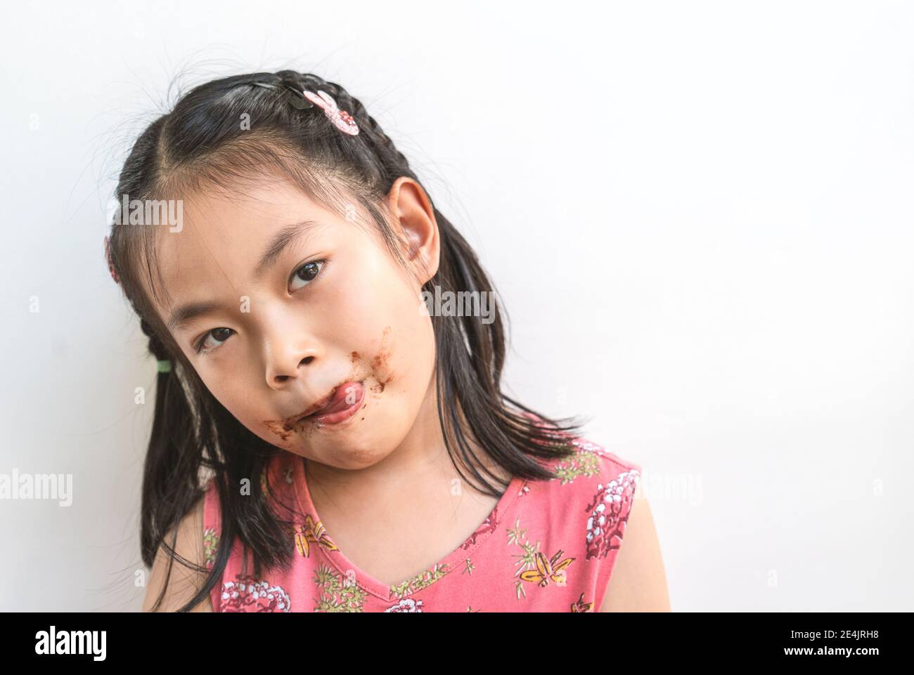 Portrait Asian nettes Kind Mädchen mit chaotisch von Schokolade um den Mund, Zunge leckt Lippen, schwarze Haare, tragen schöne rosa Kleid, isoliert Bild Stockfoto