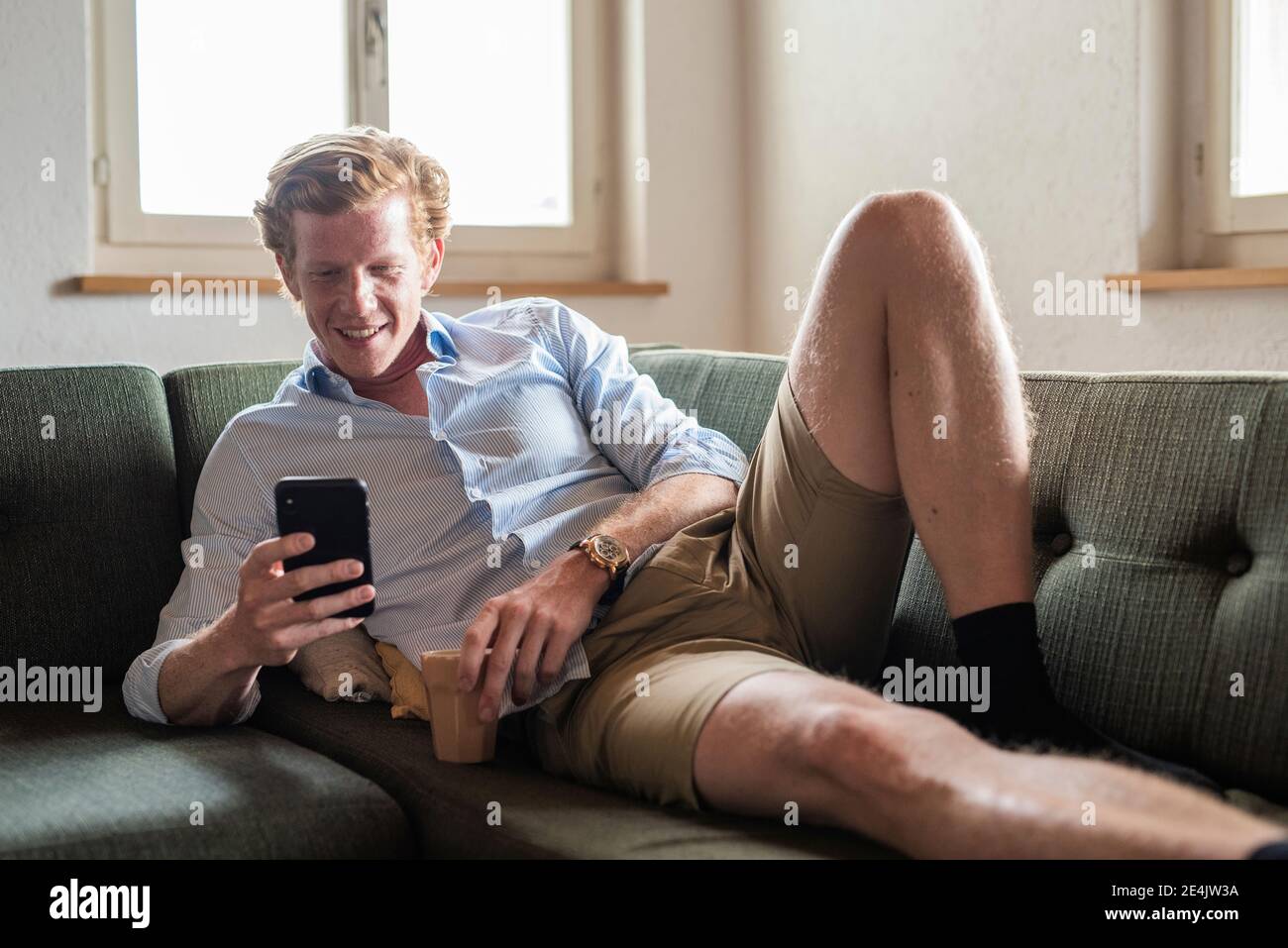 Lächelnder Mann, der Smartphone benutzt, während er auf dem Sofa liegt Wohnzimmer Stockfoto