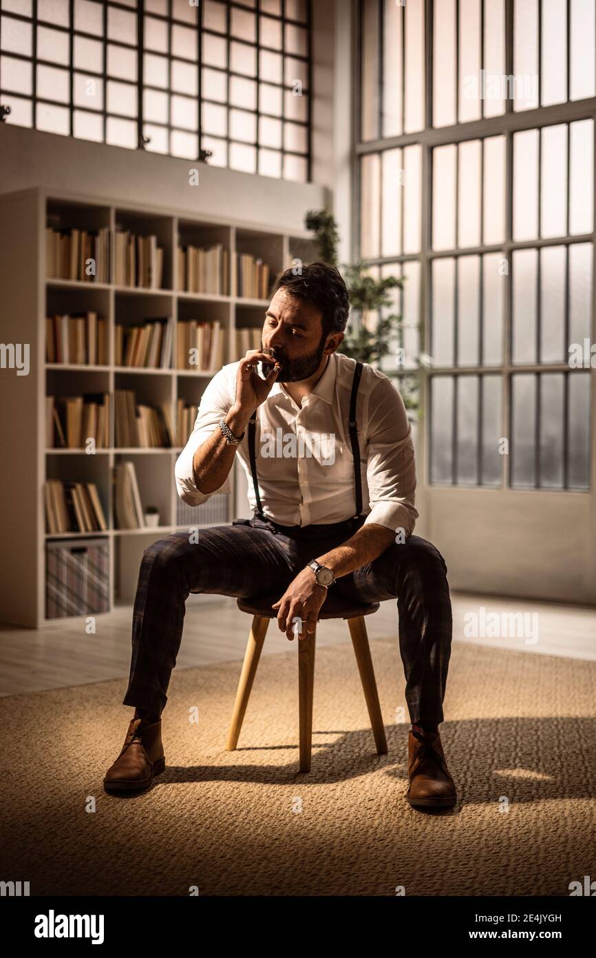 Porträt eines bärtigen Mannes, der auf einem Hocker sitzt und Zigarre raucht Stockfoto