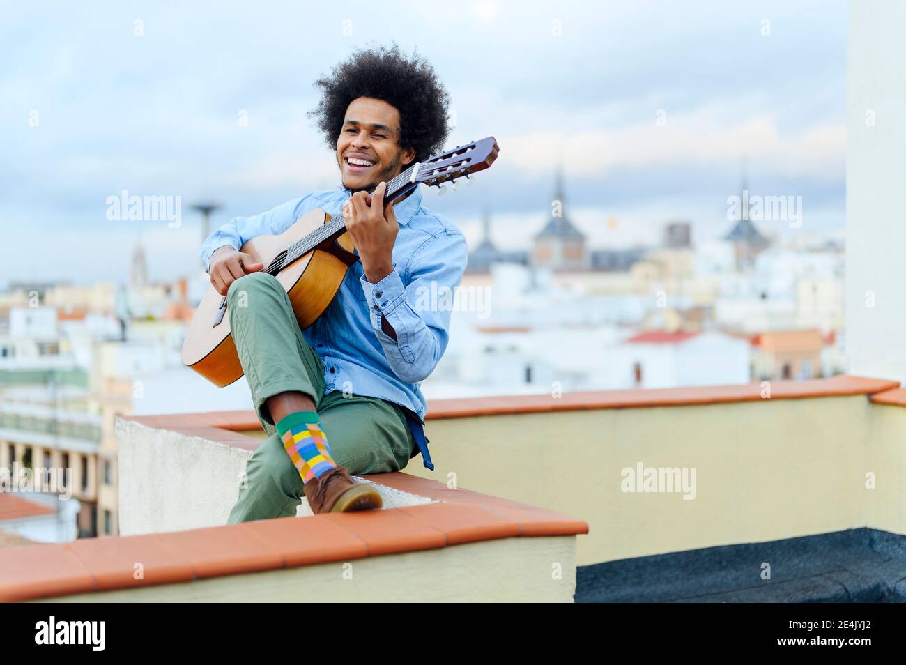 Fröhlicher junger Mann, der Gitarre spielt, während er auf der Terrasse gegen den Himmel sitzt Stockfoto