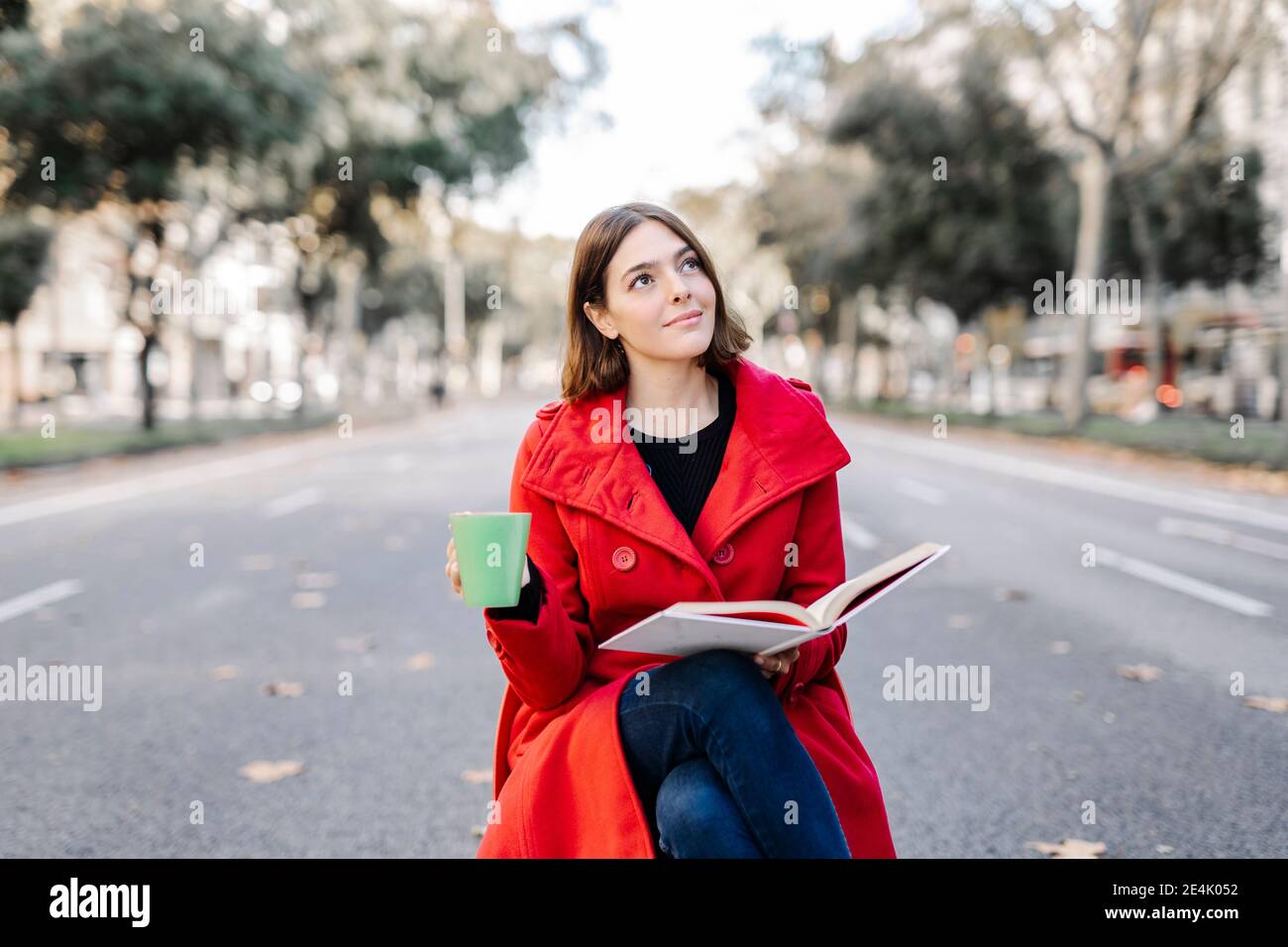 Junge Frau in roter Jacke mit Kaffeetasse Tag träumen Beim Halten des Buches auf der Straße Stockfoto