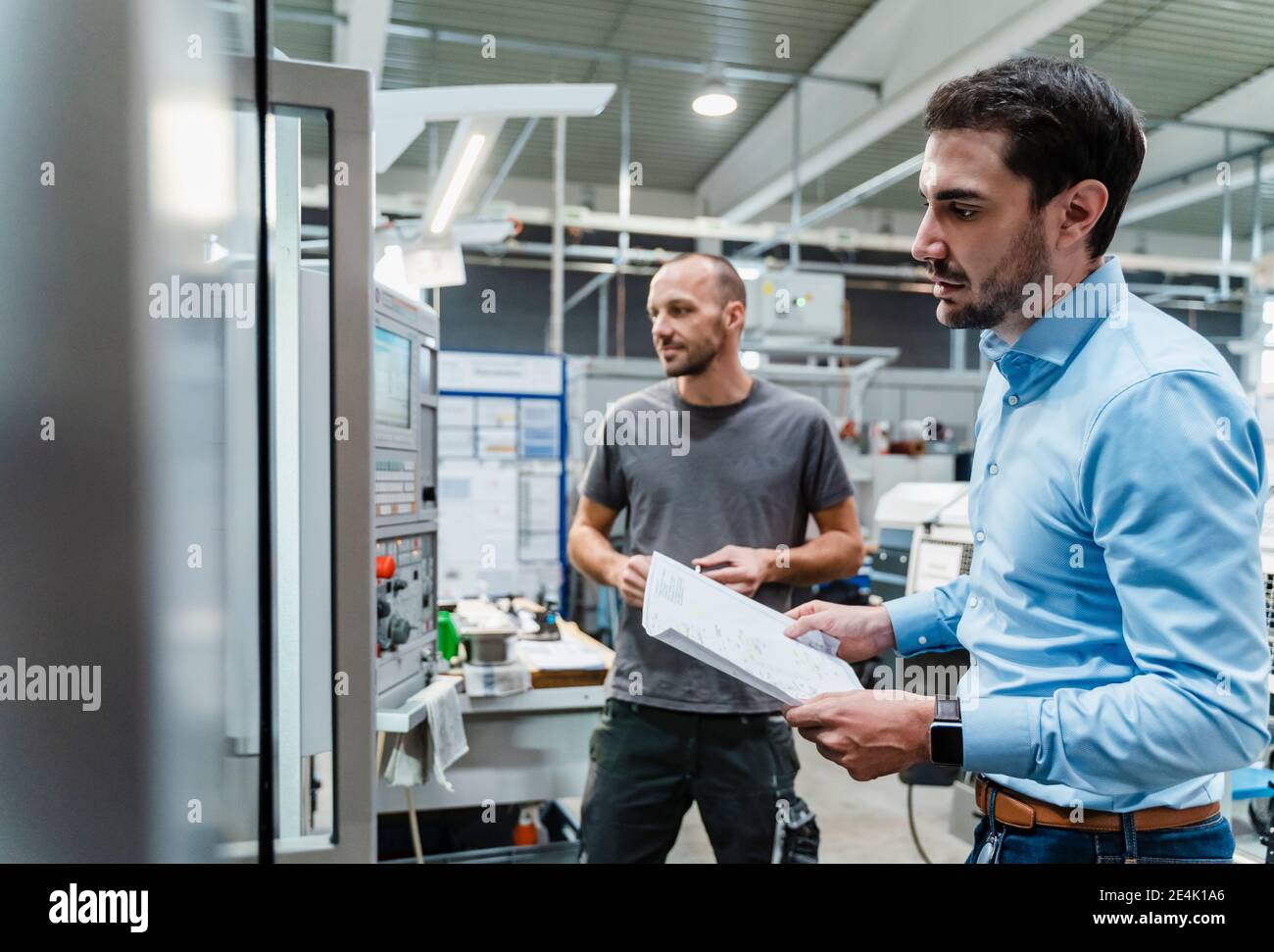 Männlicher Ingenieur mit Dokument, das Maschinen während des Stehens mit Wartung untersucht Arbeiter im Hintergrund in der Fabrik Stockfoto