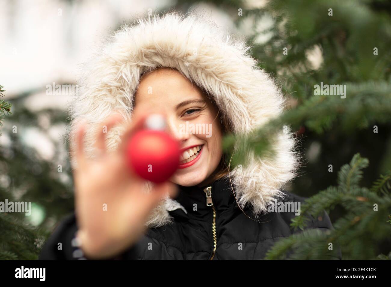 Lächelnde junge Frau trägt Pelzjacke halten Weihnachtsdekoration in der Nähe Baum Stockfoto