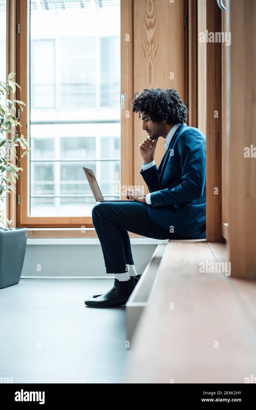 Junger Geschäftsmann mit Laptop, während er am Arbeitsplatz am Fenster sitzt Stockfoto