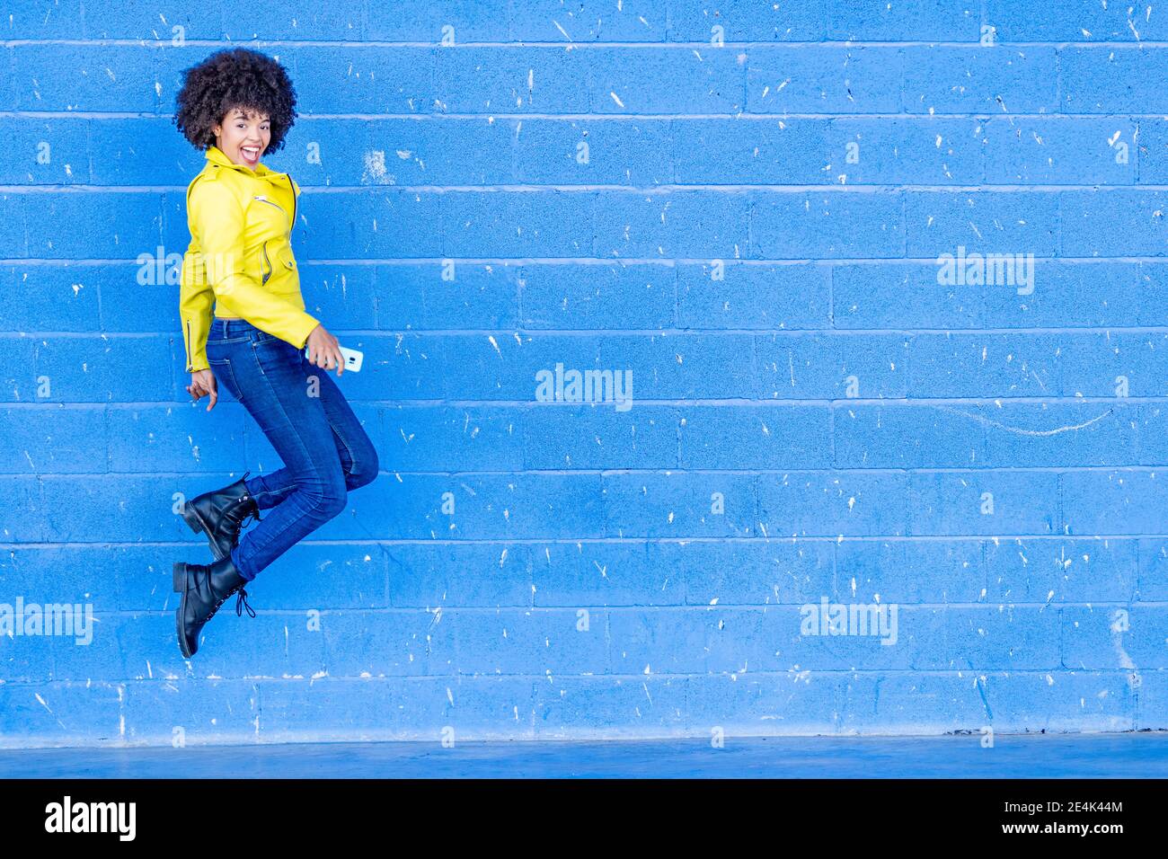 Glückliche modische Frau mit Smartphone springen gegen blaue Wand Stockfoto