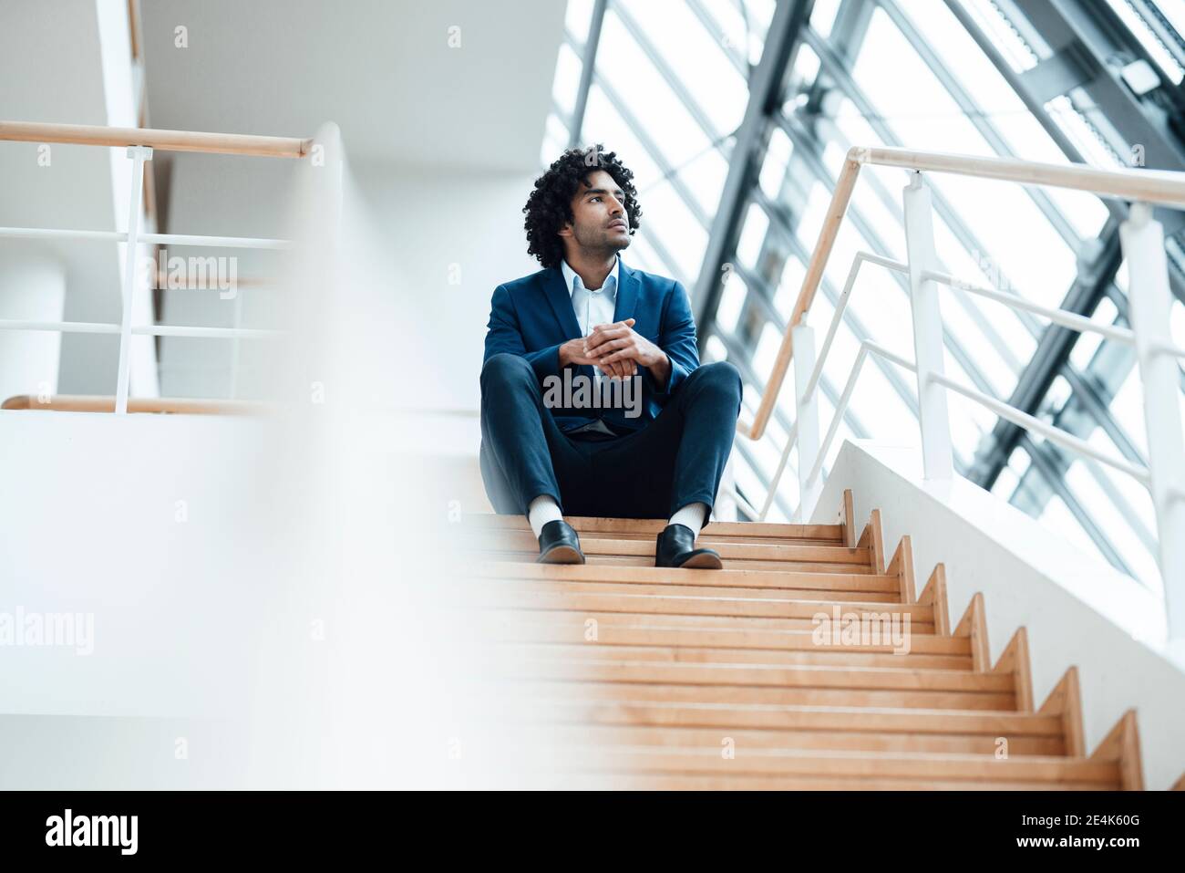 Nachdenklicher männlicher Unternehmer schaut weg, während er mit Händen zusammenhält Auf der Treppe im Büro Stockfoto