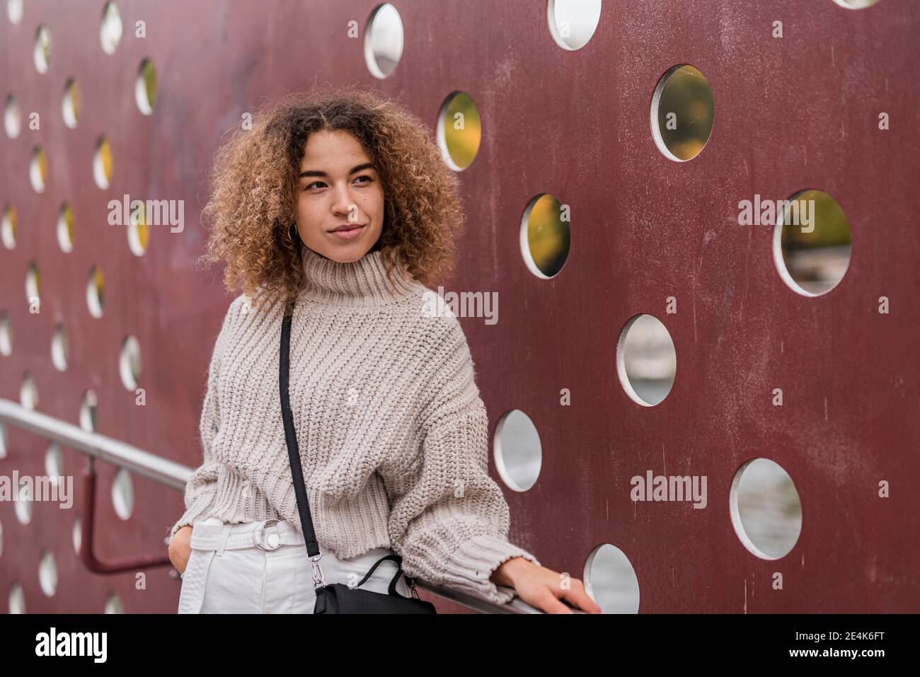 Nachdenkliche junge blonde Frau, die sich an einem Geländer lehnt, während sie gegen eine metallische Wand steht Stockfoto