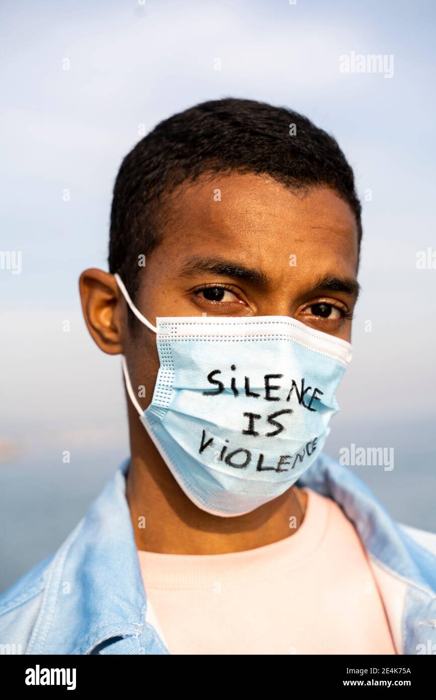 Junger Mann, der mit einer Gesichtsmaske mit der Aufschrift „Silence is Violence“ starrt Im Freien stehen Stockfoto