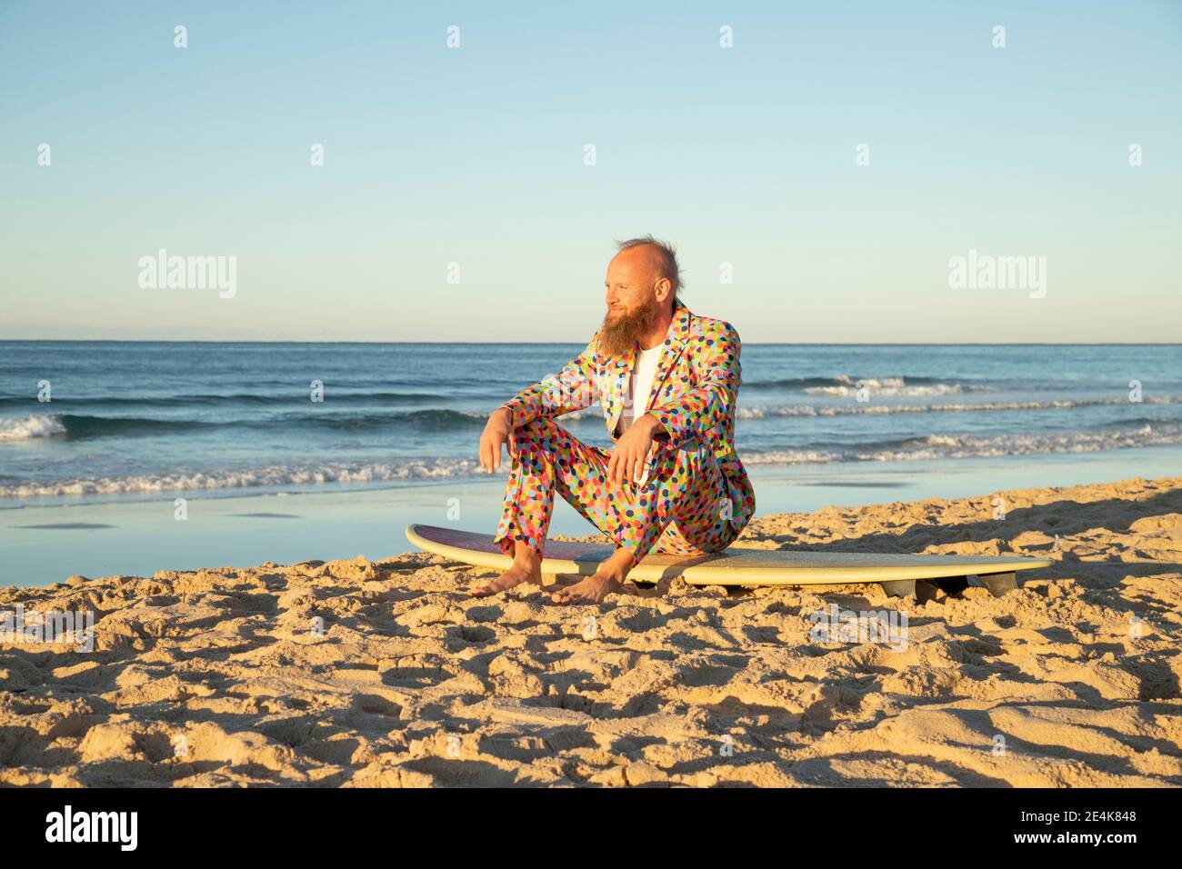Mann in farbenfrohem Anzug mit Blick auf das Meer während des Sitzens Am Surfbrett am Strand Stockfoto