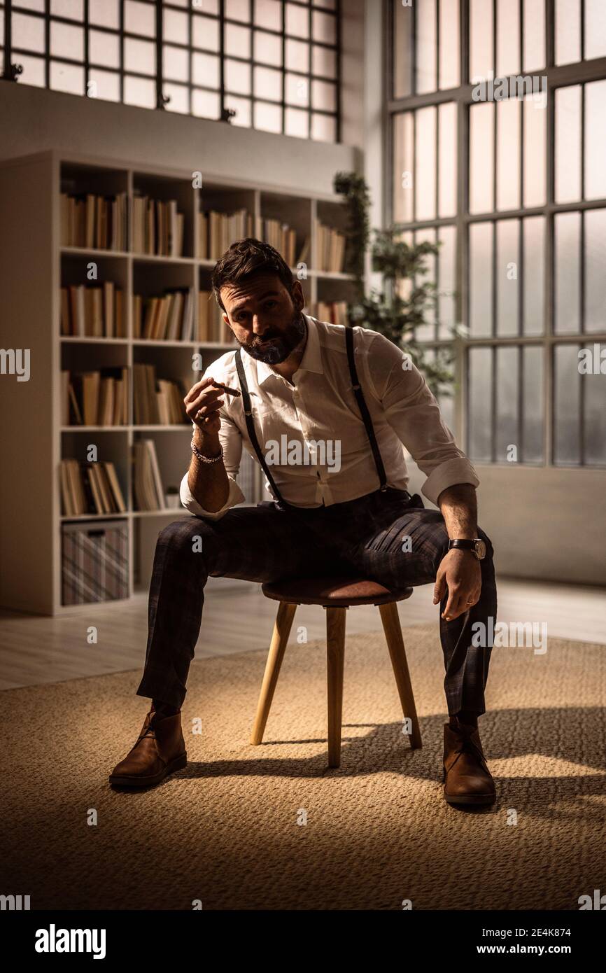 Porträt eines bärtigen Mannes, der auf einem Hocker sitzt und Zigarre raucht Stockfoto