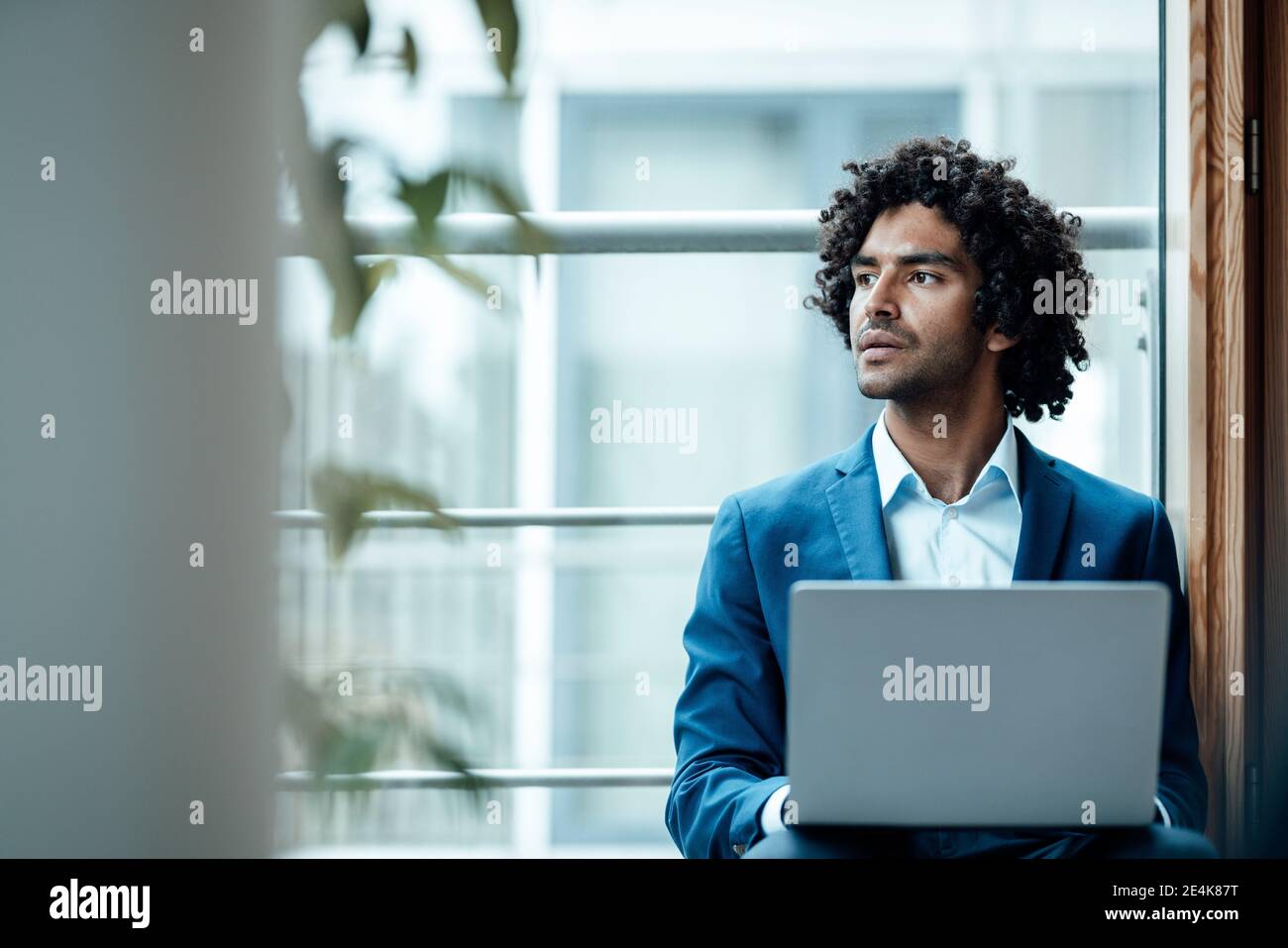 Nachdenklicher männlicher Profi schaut weg, während er mit Laptop gegen sitzt Fenster am Arbeitsplatz Stockfoto