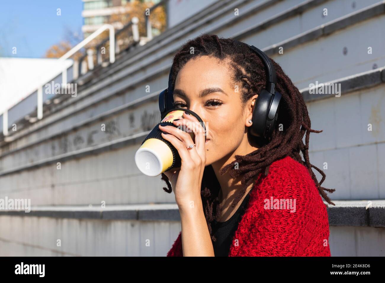 Junge Frau, die Musik hört, während sie Kaffee aus einer wiederverwendbaren Tasse trinkt An sonnigen Tag Stockfoto