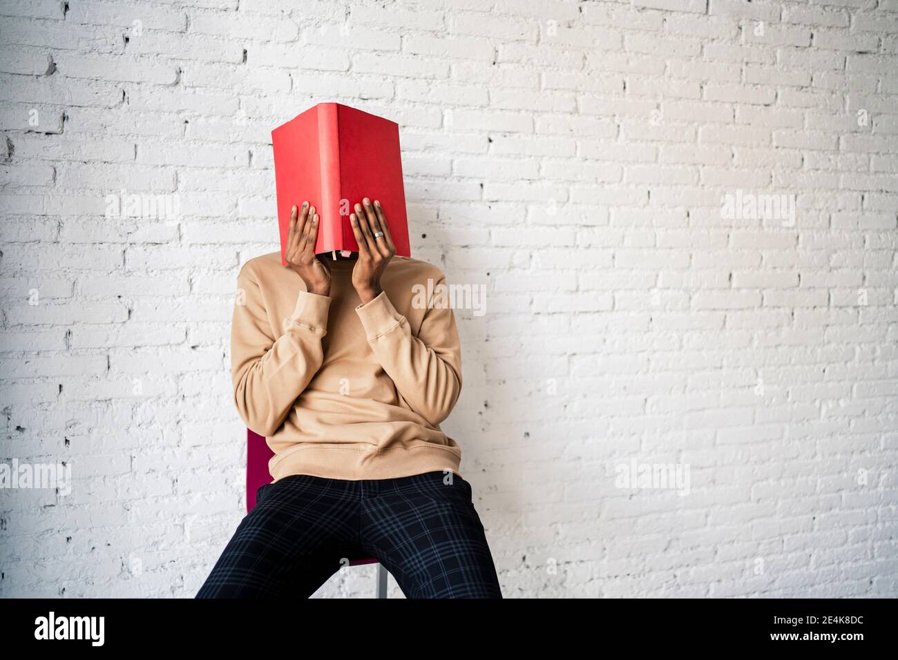 Mann bedeckt Gesicht mit rotem Buch, während er auf einem Stuhl sitzt Gegen weiße Backsteinmauer Stockfoto