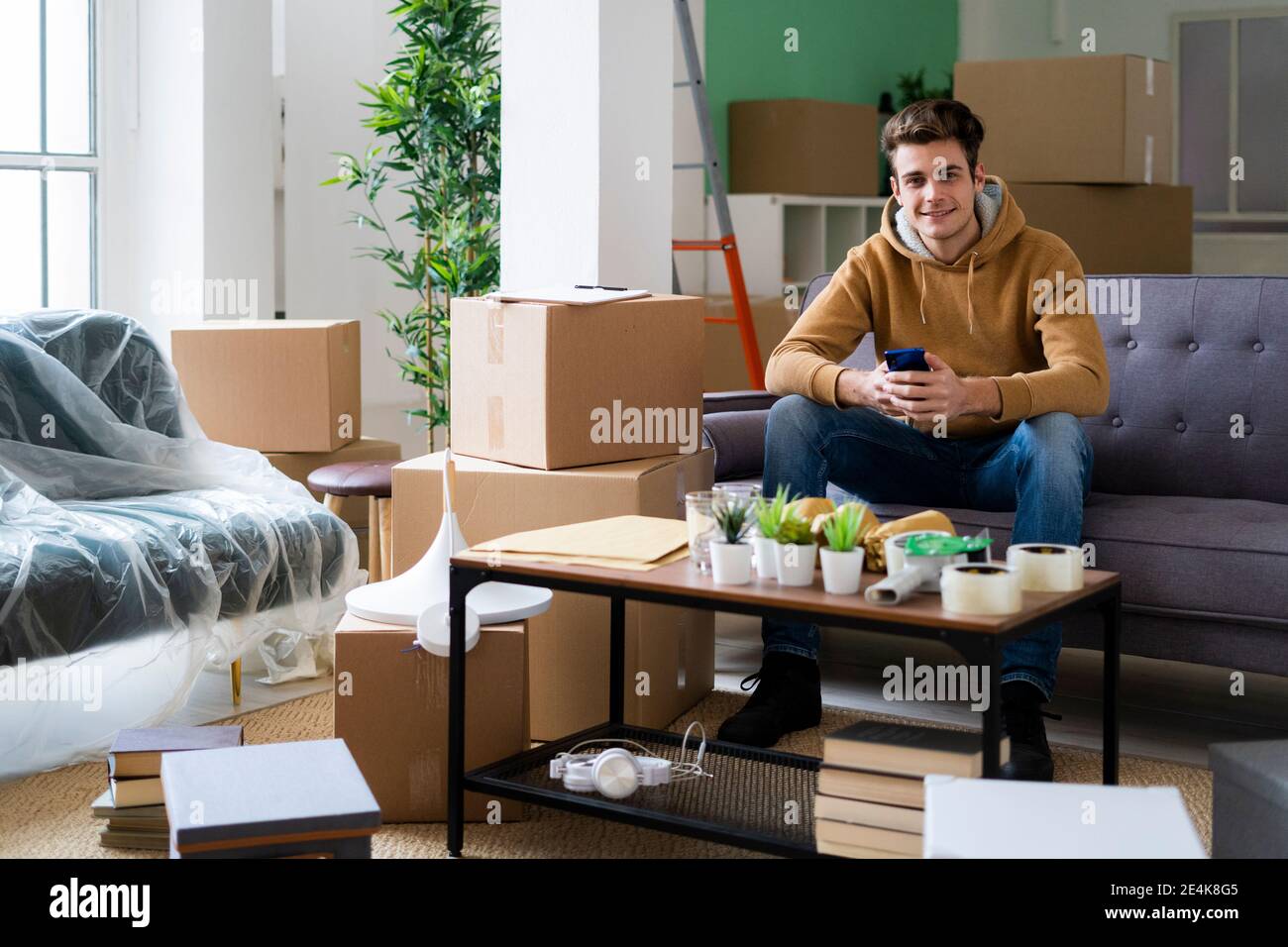 Lächelnder junger Mann mit Smartphone, der voll im Zimmer sitzt Der Kisten während des Umzugs Stockfoto