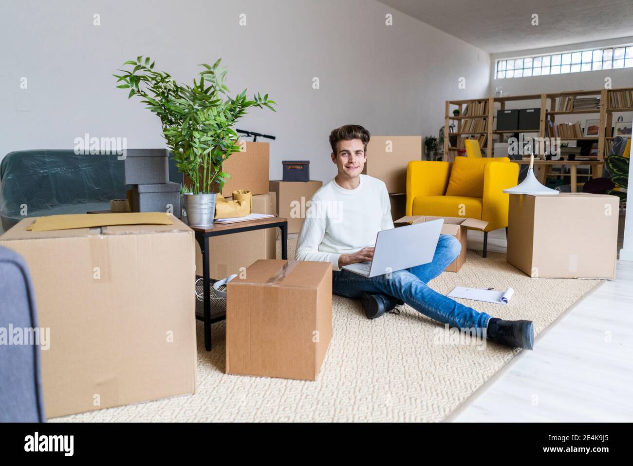 Lächelnder junger Mann mit Laptop sitzt in einem chaotischen Wohnzimmer Bei der neuen Wohnung Stockfoto