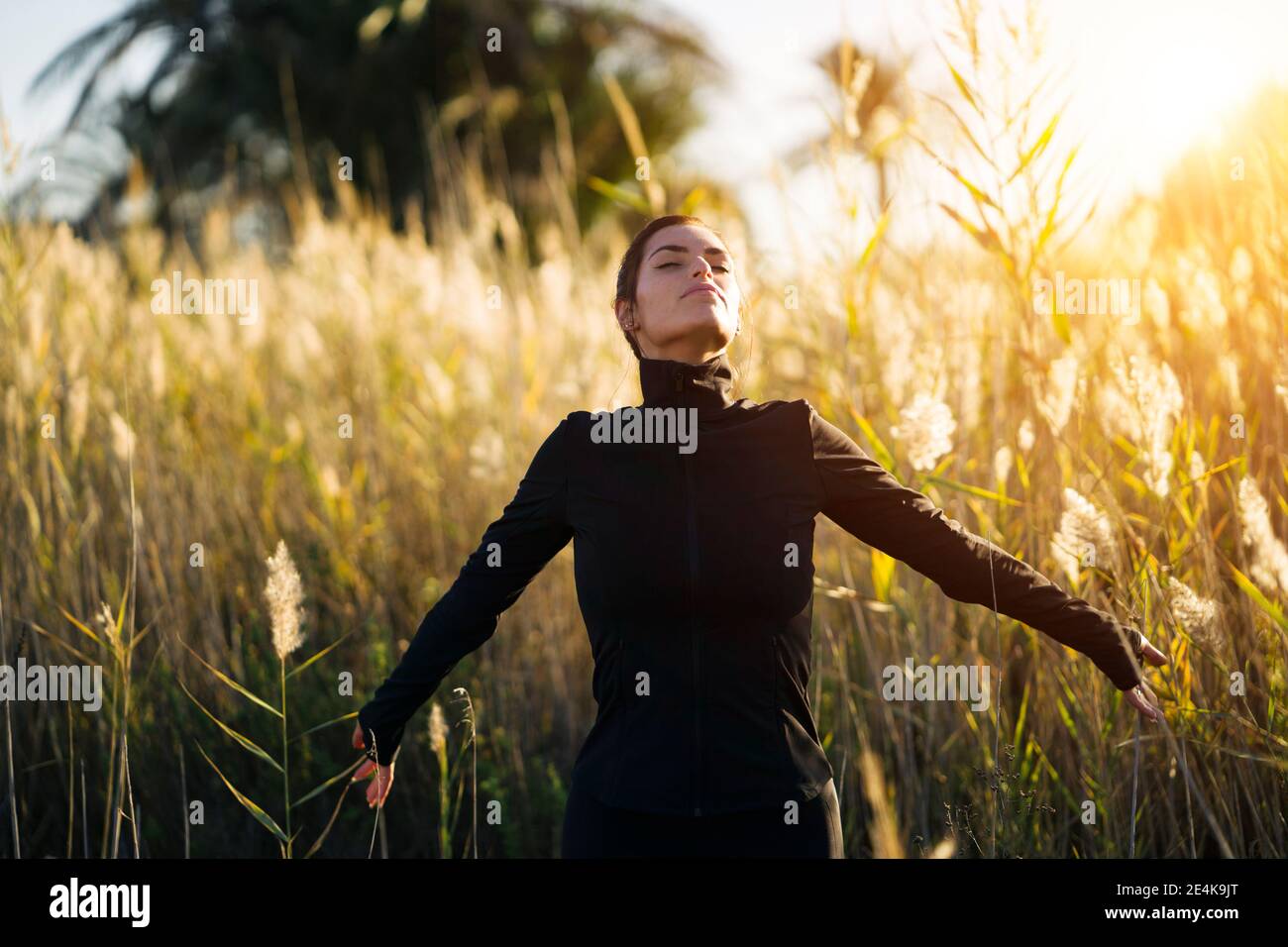 Junge Frau mit geschlossenen Augen, die während des Sonnenuntergangs gegen Pflanzen steht Stockfoto
