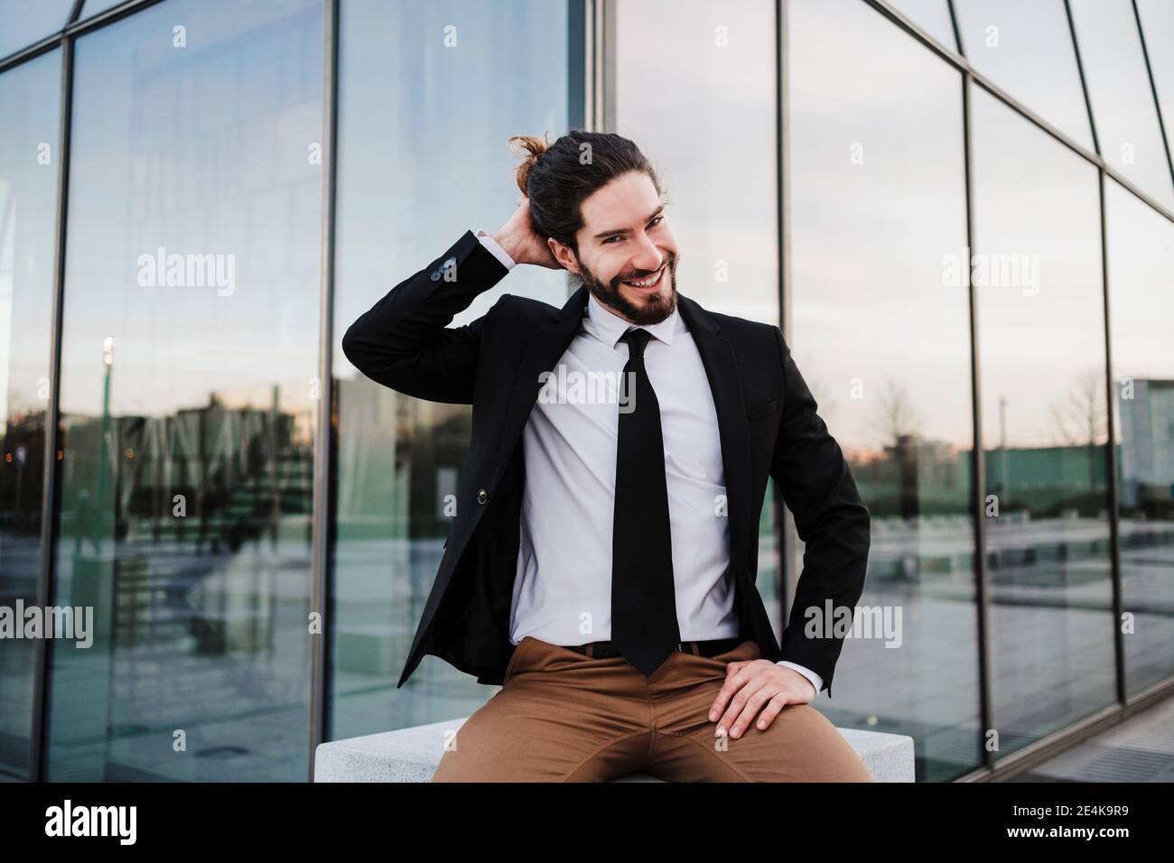 Fröhlicher Geschäftsmann mit der Hand hinter dem Kopf, der gegen das Gebäude sitzt Stockfoto