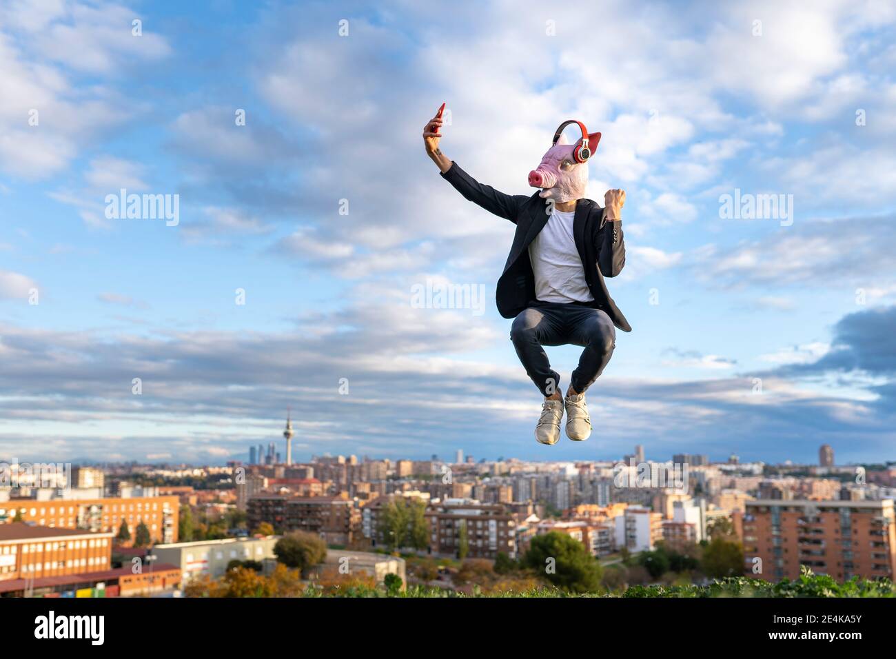 Männliche Unternehmer tragen Schwein Maske unter Selfie von Smartphone Beim Springen in der Stadt gegen den Himmel Stockfoto