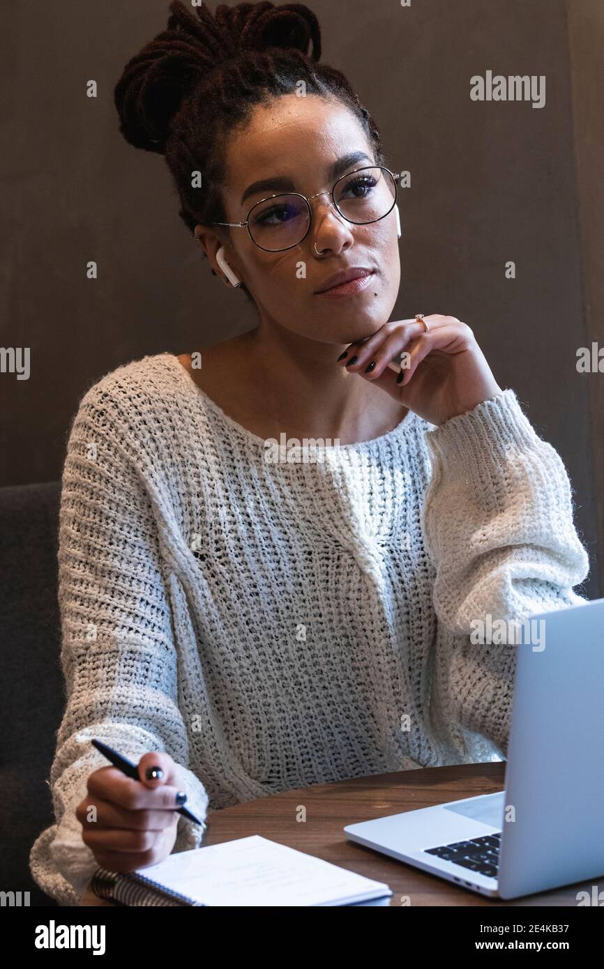 Nachdenkliche junge Frau, die im Tagebuch schreibt, während sie mit einem Laptop sitzt Im Café Stockfoto