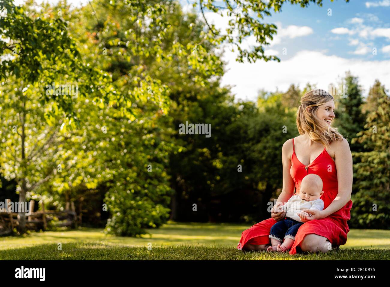 Lächelnde Mutter hält den kleinen Jungen, während sie auf Gras kniet Im Park Stockfoto