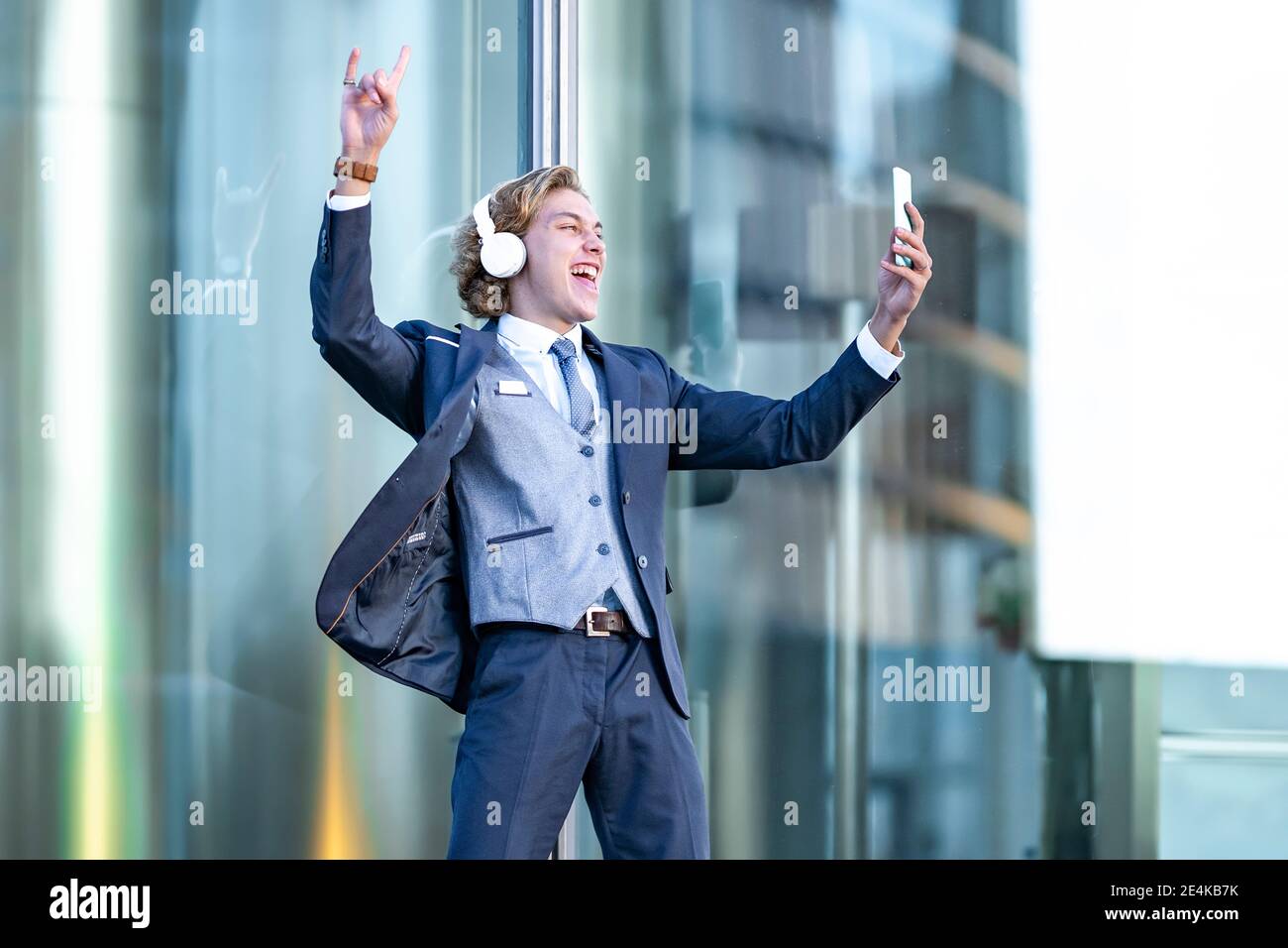 Fröhlicher junger Geschäftsmann, der Musik hört, während er Selfie gegen Glas nimmt Stockfoto