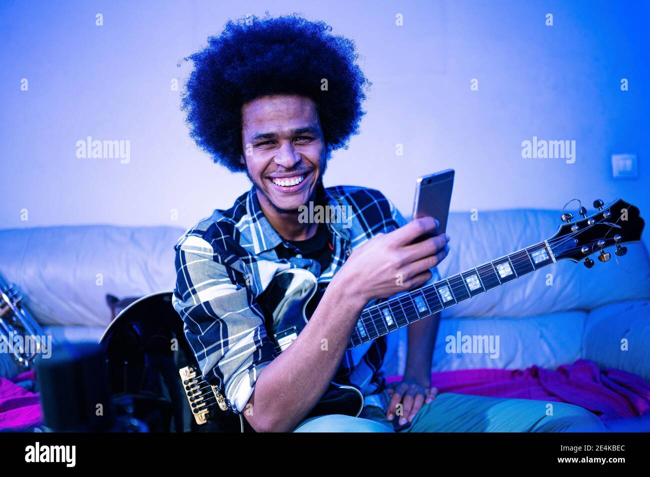 Glücklicher Afro-Mann mit Gitarre, der wegschaute, während er das Mobiltelefon im blauen Raum hielt Stockfoto