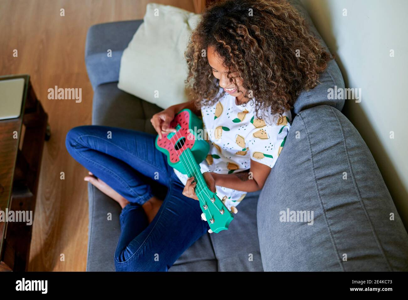 Lächelnde junge Frau mit lockigen Haaren üben Ukulele zu spielen Im Wohnzimmer Stockfoto