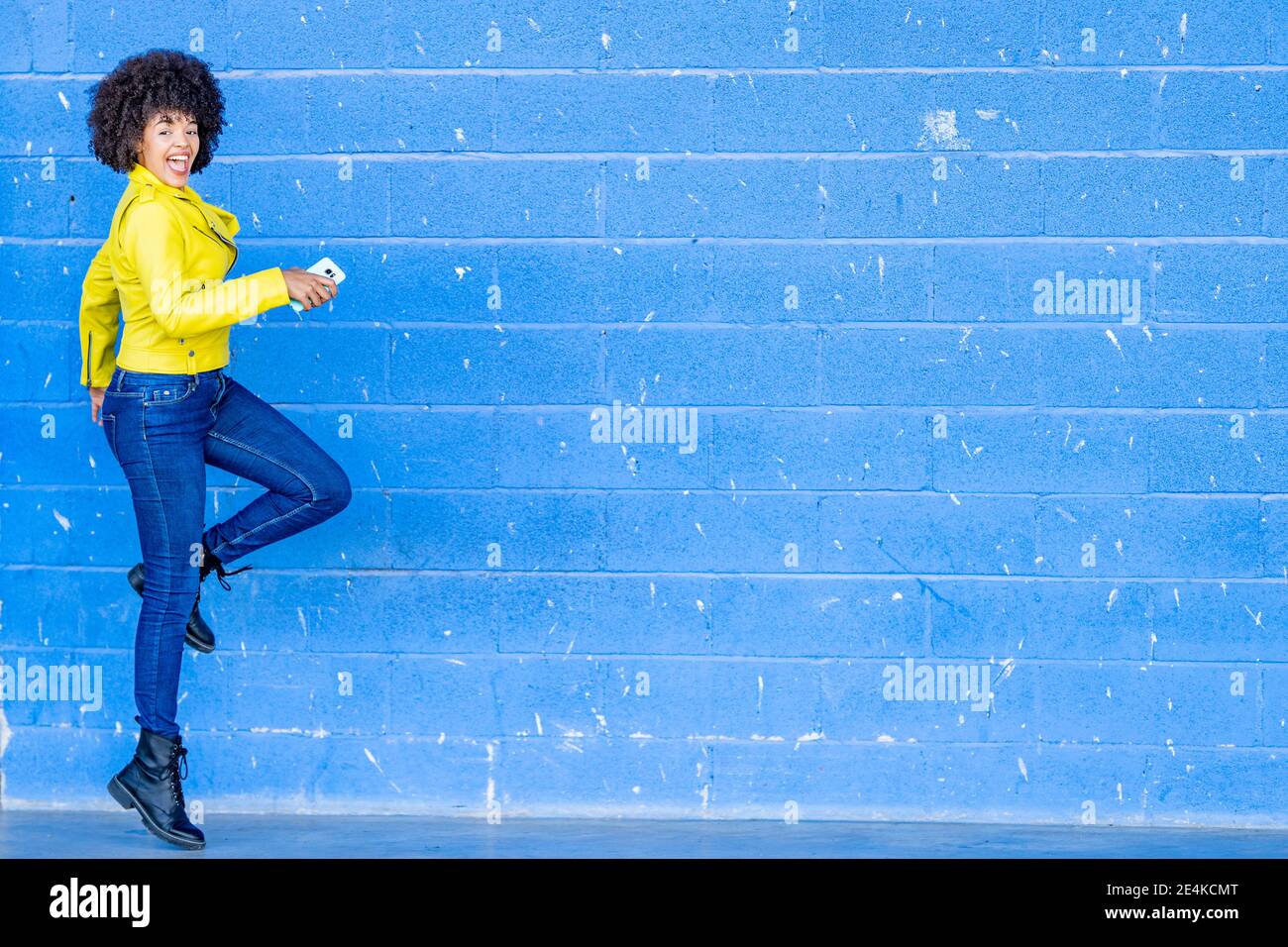 Fröhliche junge Frau mit Handy, das an der blauen Wand tanzt Stockfoto