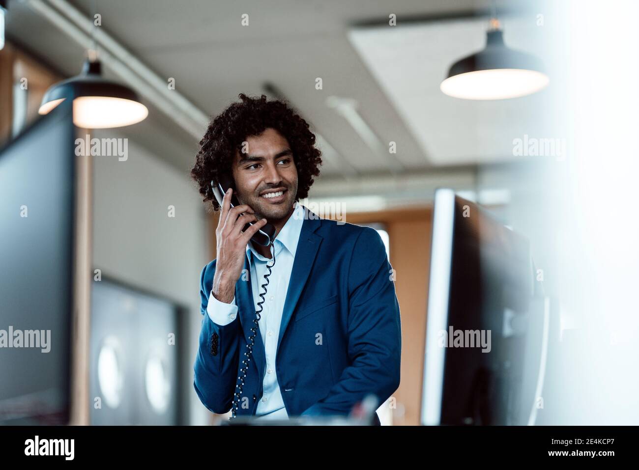 Lächelnder junger Geschäftsmann, der am Telefon spricht und dabei wegschaut Arbeitsplatz Stockfoto