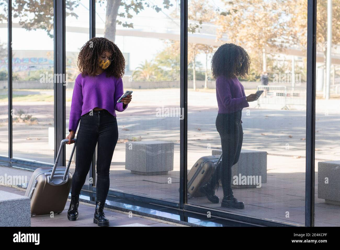 Junge Frau in schützende Gesichtsmaske mit Smartphone, während Spaziergang mit Gepäck am Glas Stockfoto